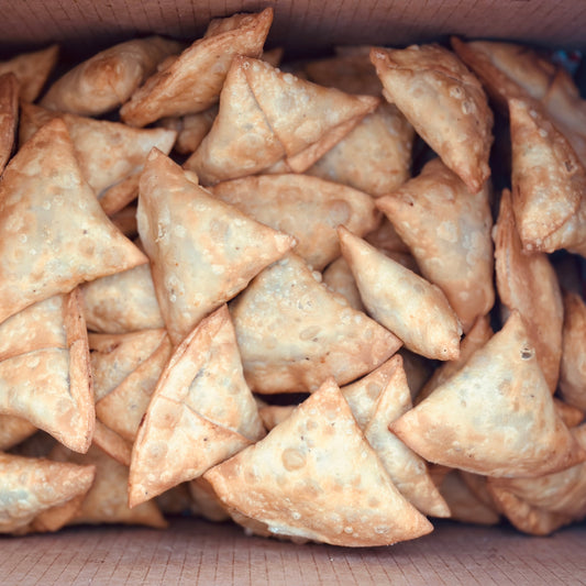 A golden, flaky handmade potato and peas samosa served on a vintage bone-china plate, accompanied by a container of chutney.