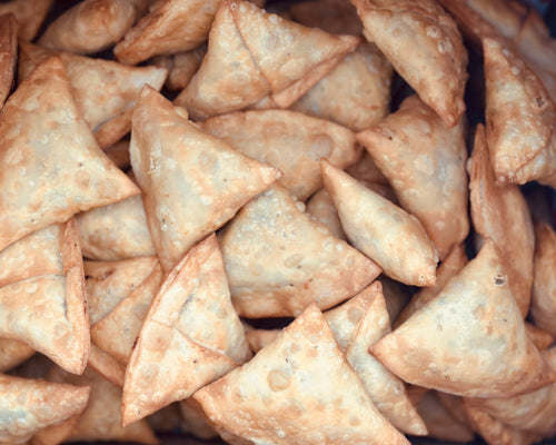 A golden, flaky handmade potato and peas samosa served on a vintage bone-china plate, accompanied by a container of chutney.