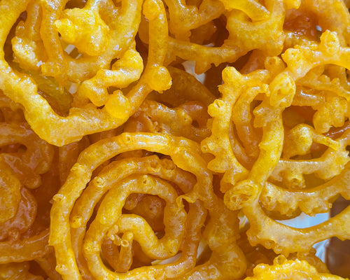 Crisp, golden Jalebis glistening with cardamom syrup, elegantly presented on a tiered silver cake stand alongside a vintage teacup.