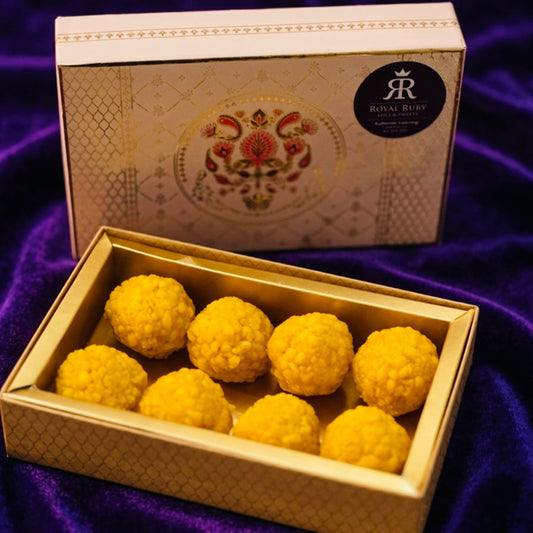 A golden-yellow box filled with 12 round, handcrafted Besan Ladoos, garnished with chopped nuts, displayed on a purple velvet surface next to a Royal Ruby branded gift box.